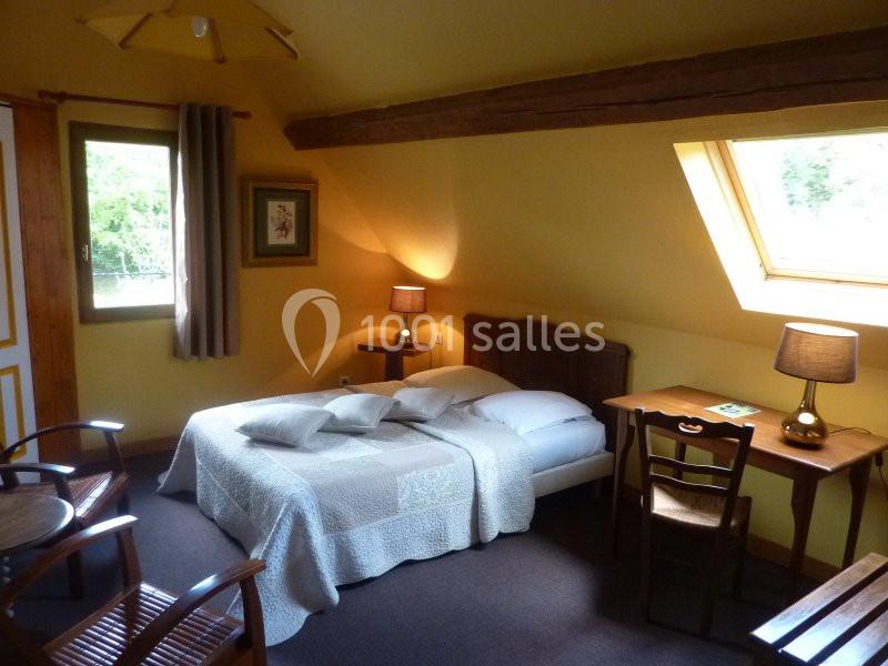 Chambre mansardée avec lit double, bureau, chaises en bois, lampes allumées et fenêtre laissant entrer la lumière naturelle.