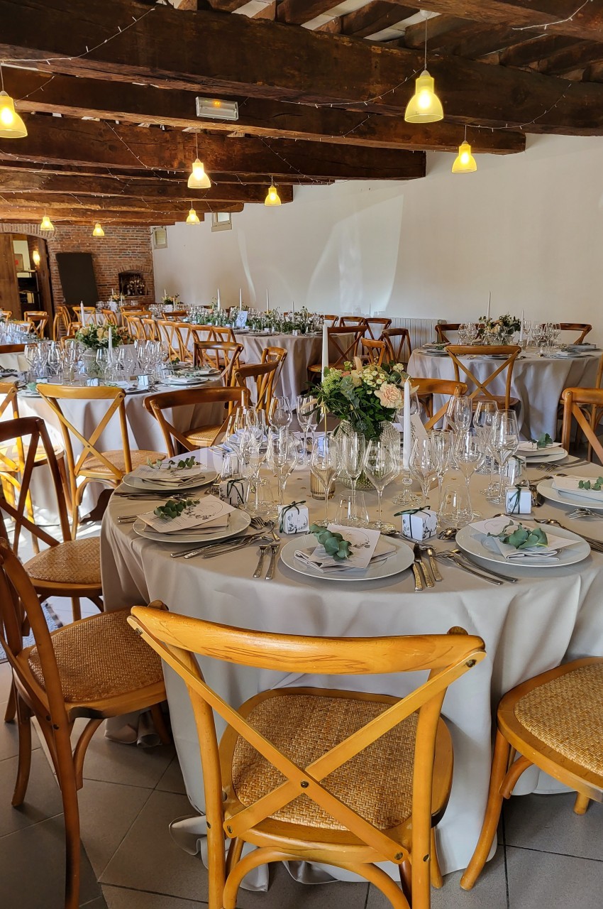 Salle de réception décorée avec des tables rondes dressées, chaises en bois et éclairage chaleureux.