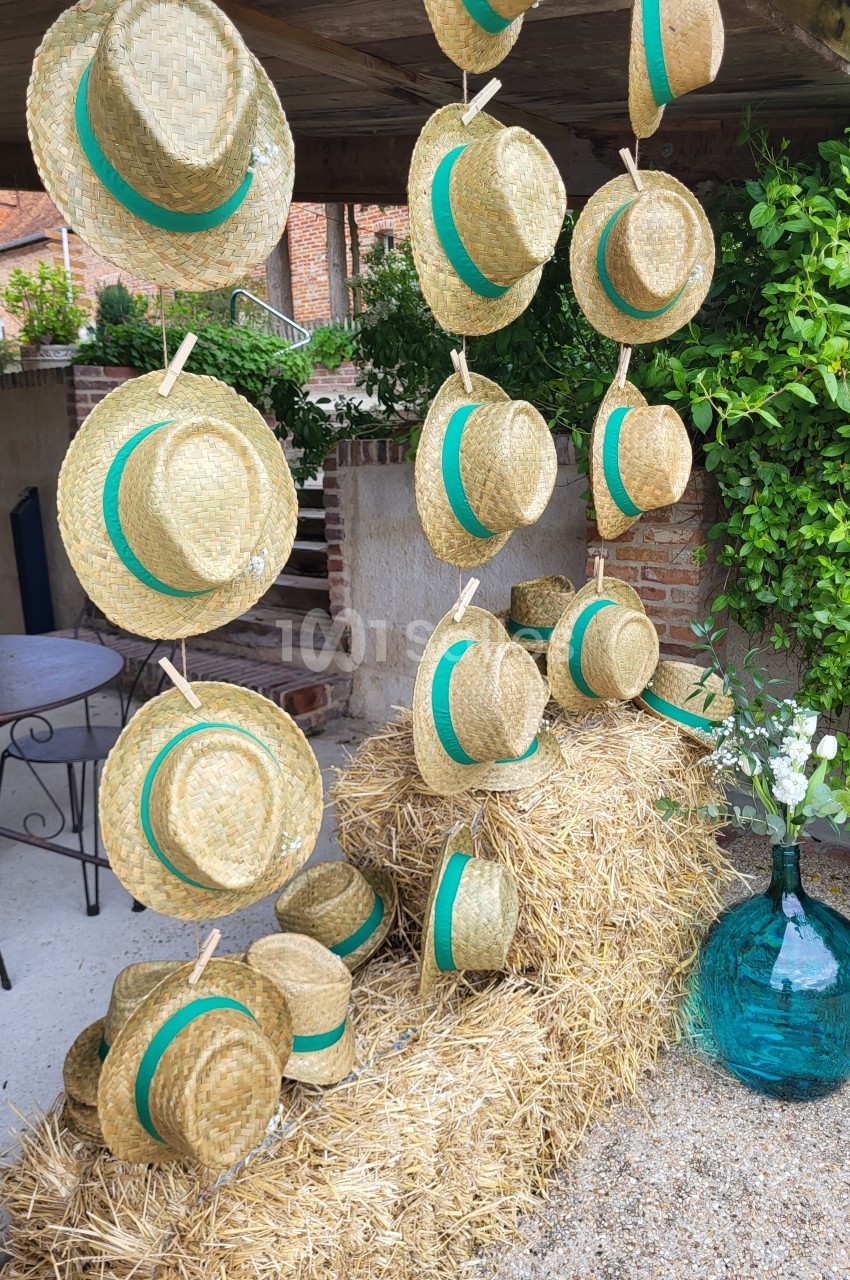 Chapeaux de paille avec rubans verts suspendus et posés sur des bottes de foin, près d'une jarre en verre turquoise.