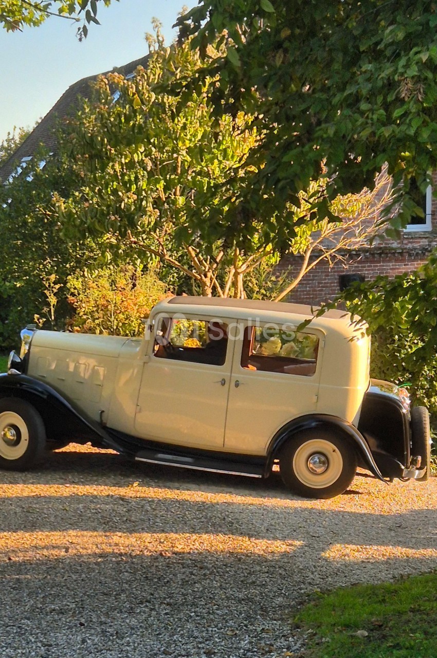 Voiture ancienne beige garée sur une allée gravillonnée, entourée de verdure et d'arbres sous une lumière douce.