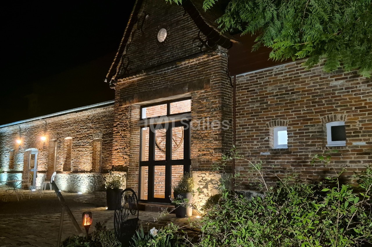 Façade en briques d'un bâtiment éclairé la nuit, entourée de végétation et d'un chemin pavé.