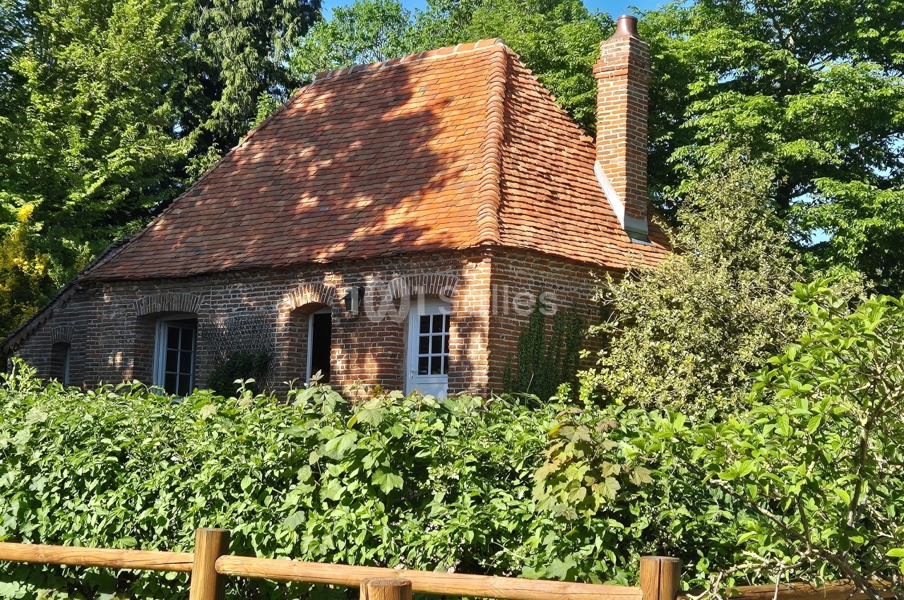 Maison en briques rouges avec toit en tuiles, entourée de verdure et d'une haie, par une journée ensoleillée.