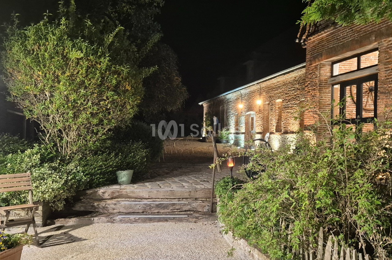 Cour extérieure éclairée de nuit, avec un bâtiment en briques, des plantes et un banc en bois.