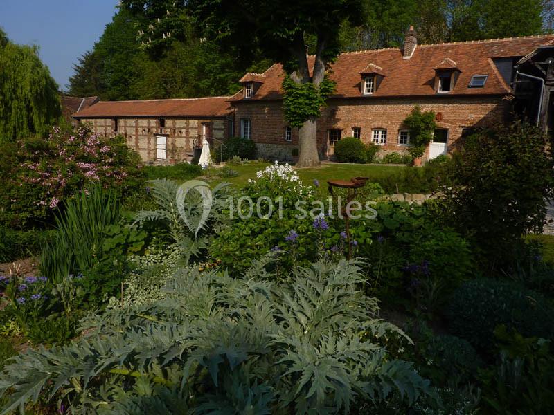 Jardin fleuri avec des plantes variées devant une maison en briques et toit en tuiles, entourée d'arbres.