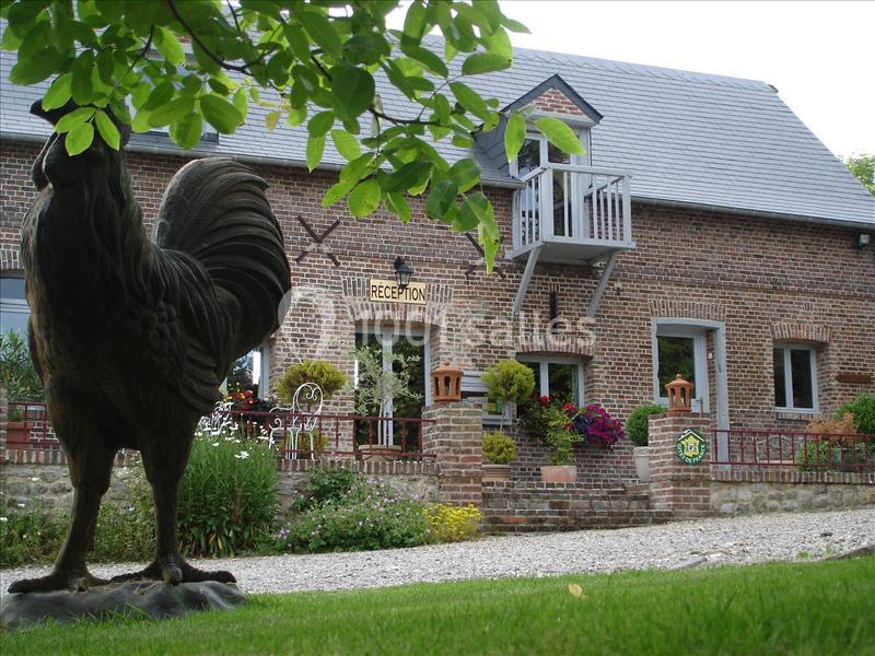 Façade en briques d'un bâtiment avec balcon, jardin fleuri et statue de coq au premier plan.