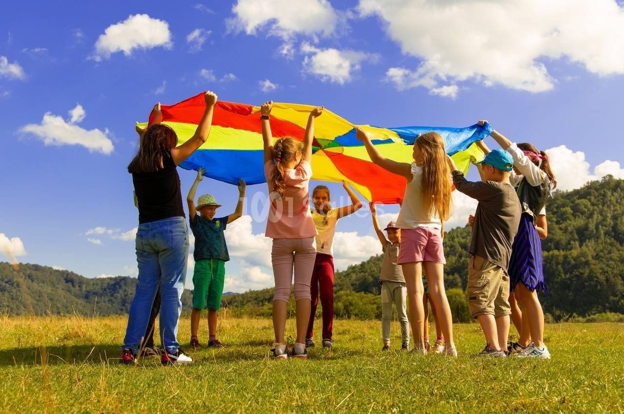 Des enfants jouent ensemble en tenant un parachute coloré dans un champ sous un ciel ensoleillé.