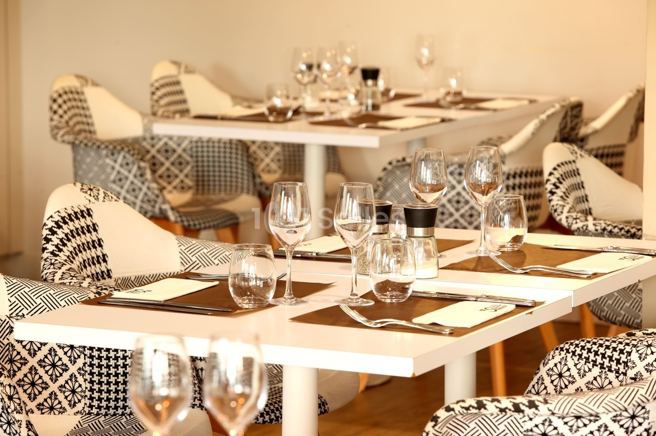 Salle de restaurant avec tables dressées, nappes blanches, verres, assiettes et chaises au motif géométrique.