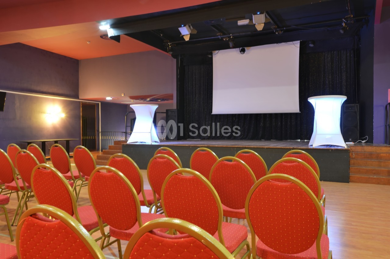 Salle de conférence avec des chaises rouges alignées face à une scène équipée d'un écran de projection.