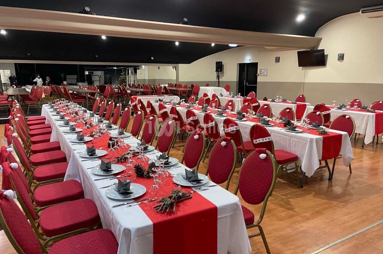 Salle de réception décorée avec des tables alignées, nappes blanches, chemins de table rouges et chaises assorties.