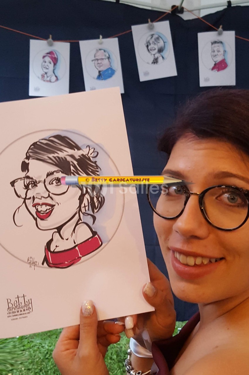 Femme souriante tenant une caricature d'elle-même, réalisée sur papier, avec d'autres dessins exposés en arrière-plan.