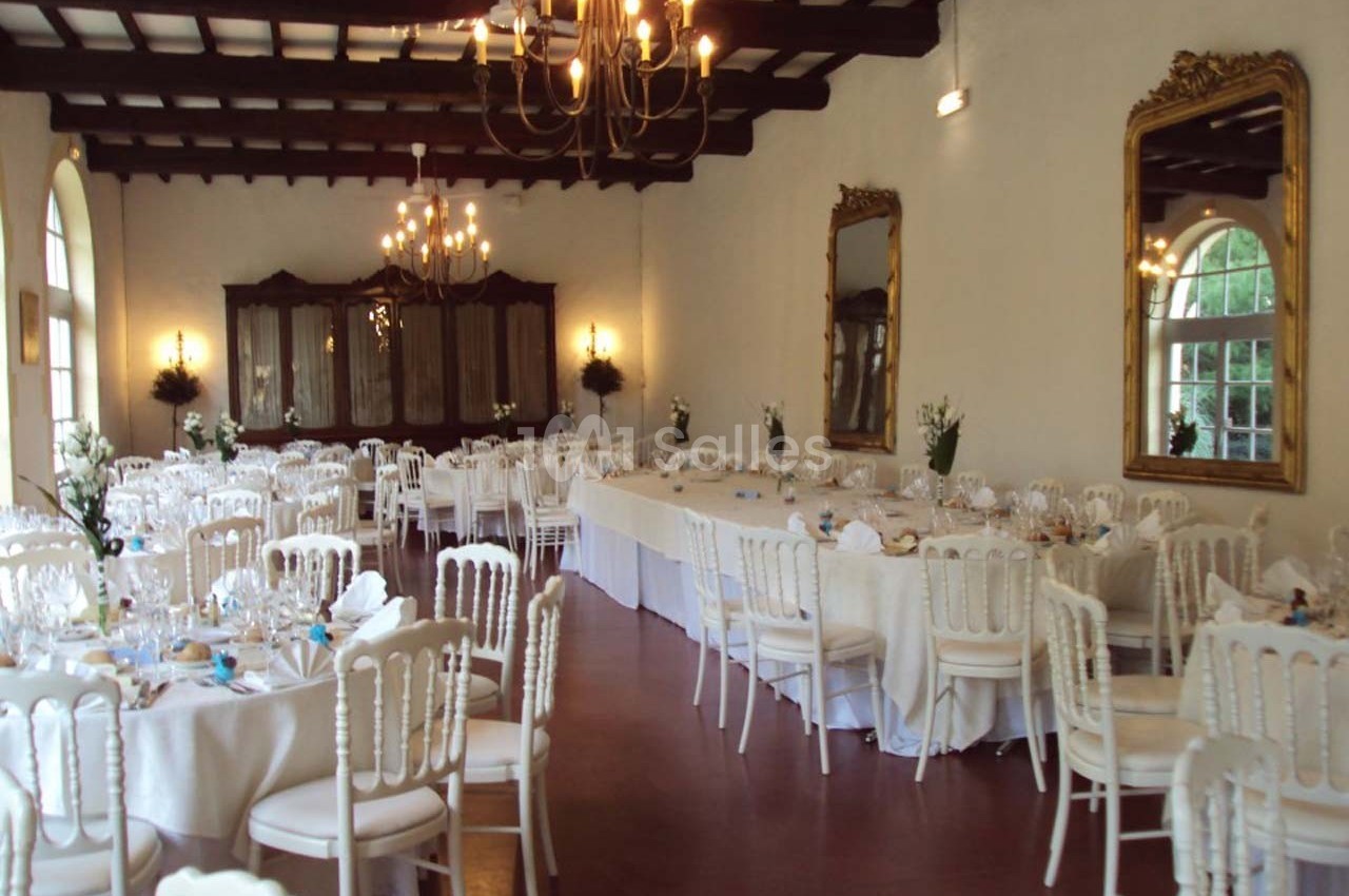 Salle de réception élégante avec tables rondes dressées, chaises blanches et lustres suspendus au plafond.