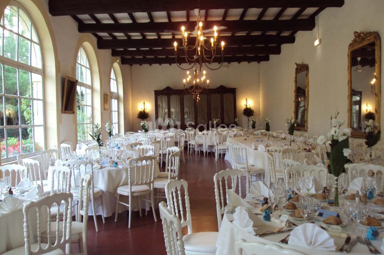 Salle de réception élégante avec tables rondes dressées, chaises blanches et grandes fenêtres laissant entrer la lumière.
