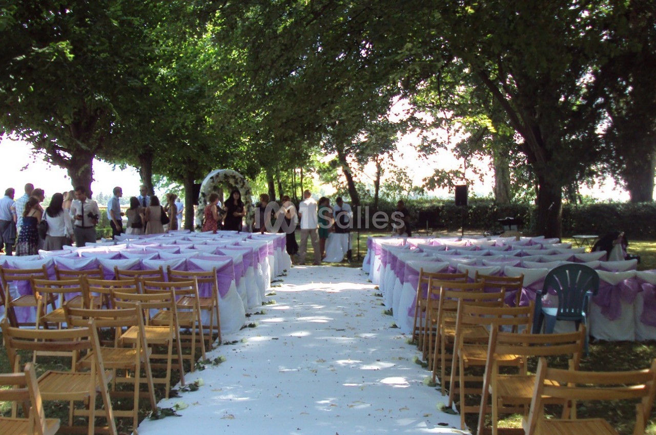 Allée centrale bordée de chaises décorées en violet, menant à une arche pour une cérémonie en plein air sous des arbres.