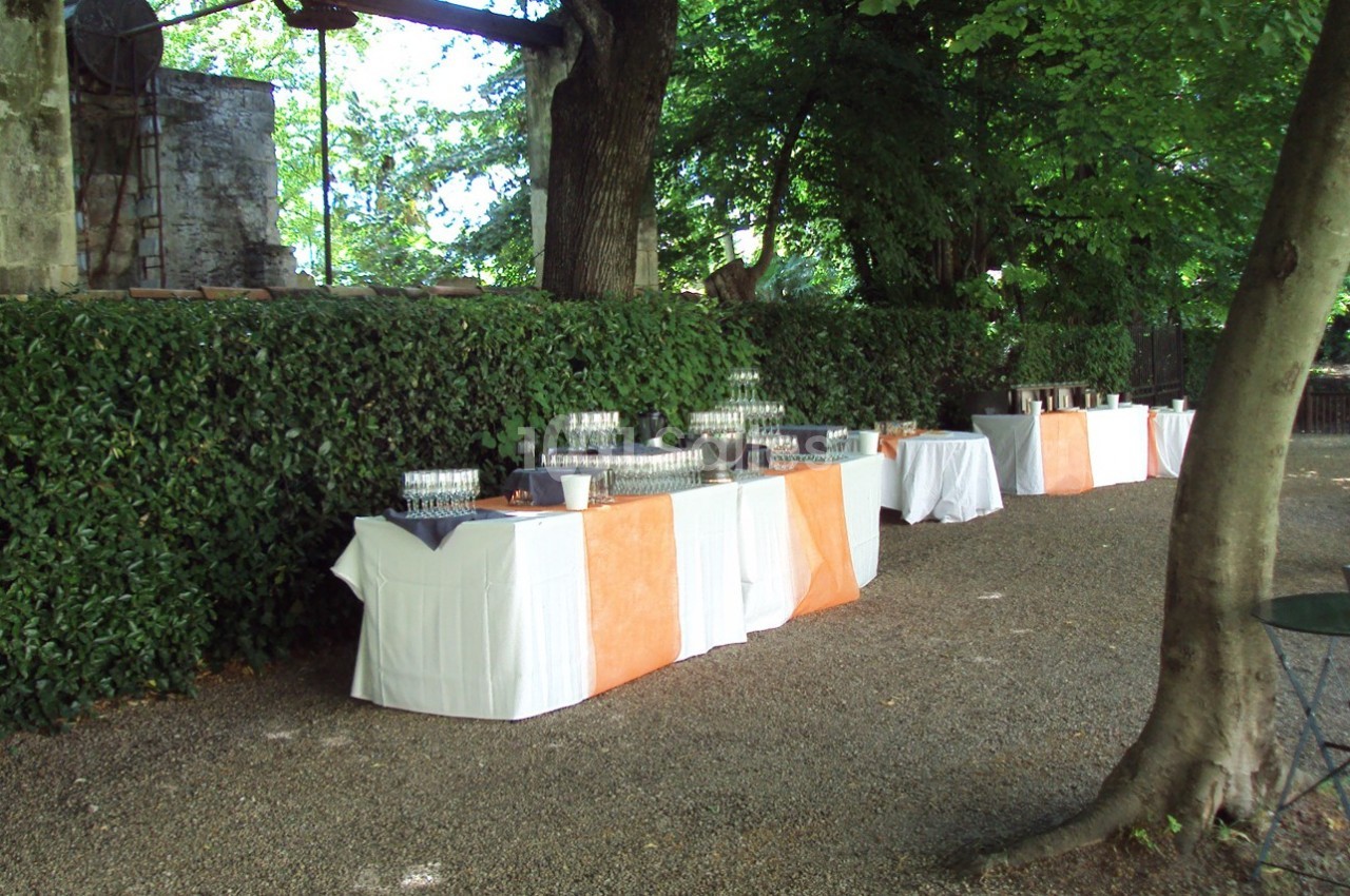 Tables dressées avec nappes blanches et oranges, disposées en extérieur près d'une haie et d'arbres, pour un événement.