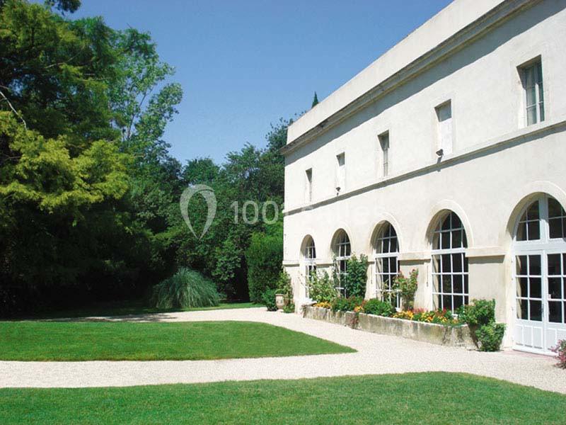 Façade d'un bâtiment ancien avec des arches vitrées, entouré d'un jardin verdoyant et d'une allée en gravier.