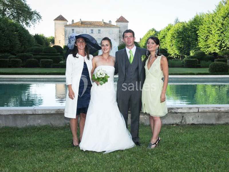 Un couple de mariés pose avec deux invités devant un bassin et un château dans un jardin verdoyant.