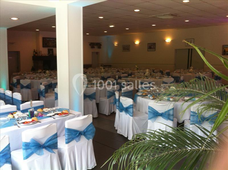 Salle de réception aménagée avec tables dressées, chaises blanches ornées de noeuds bleus et plantes vertes.
