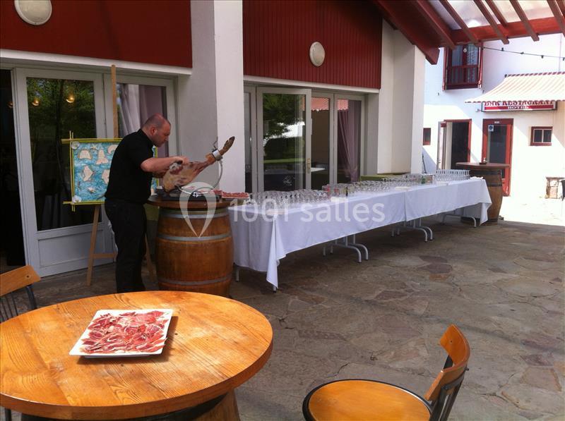 Terrasse avec tables rondes en bois, traiteur préparant des charcuteries près d'une table dressée blanche, bâtiment rouge et blanc en arrière-plan.