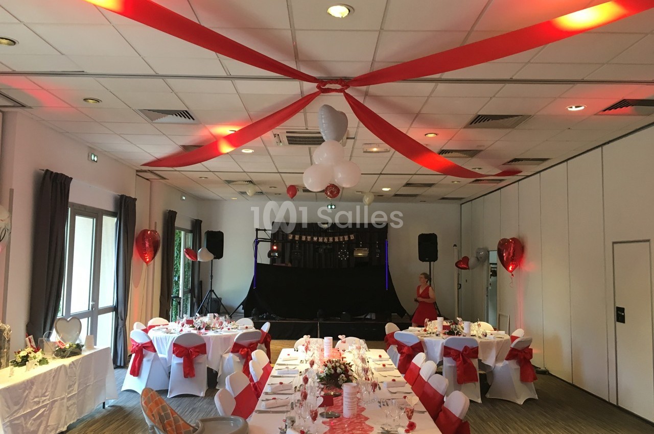 Salle décorée pour une réception avec tables dressées, nappes blanches, rubans rouges et ballons suspendus.