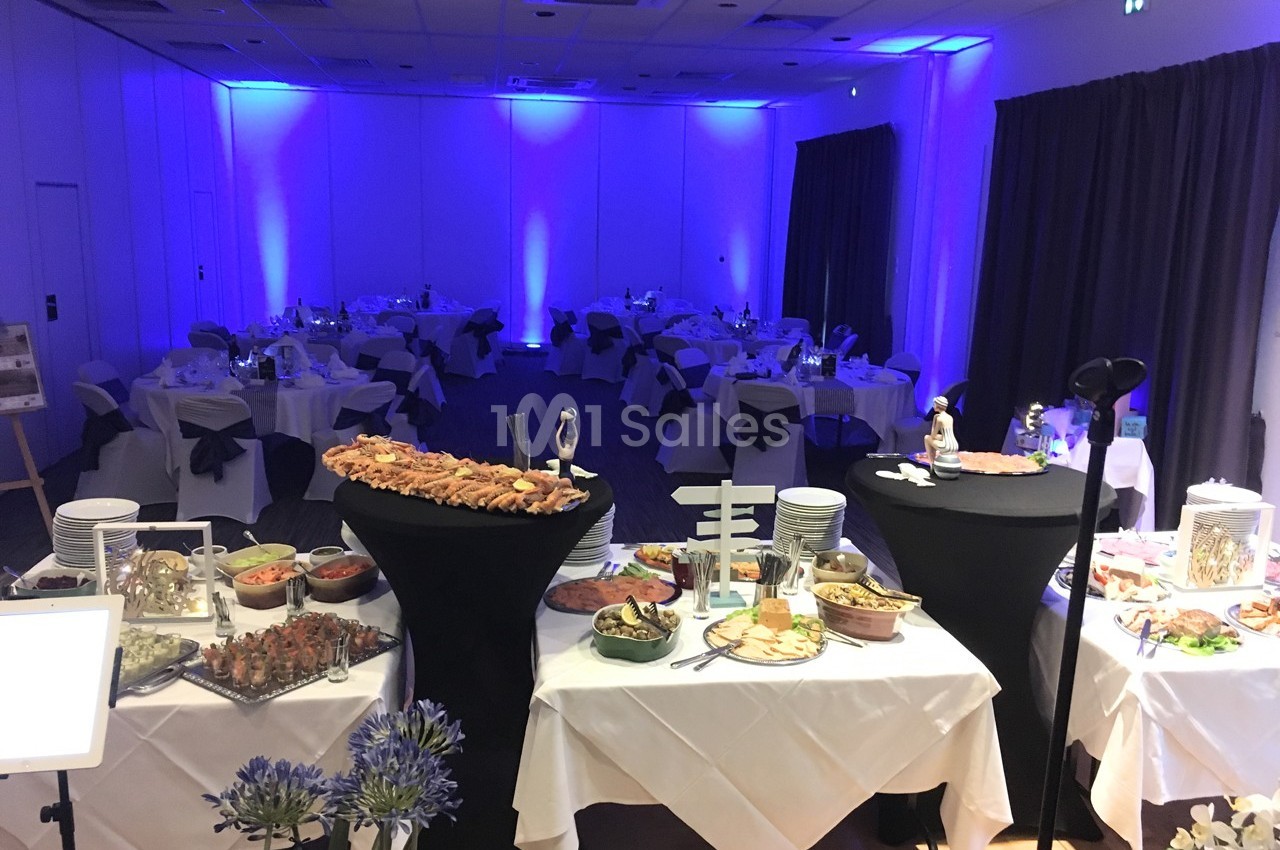 Salle de réception éclairée en bleu avec tables dressées et buffet varié au premier plan.