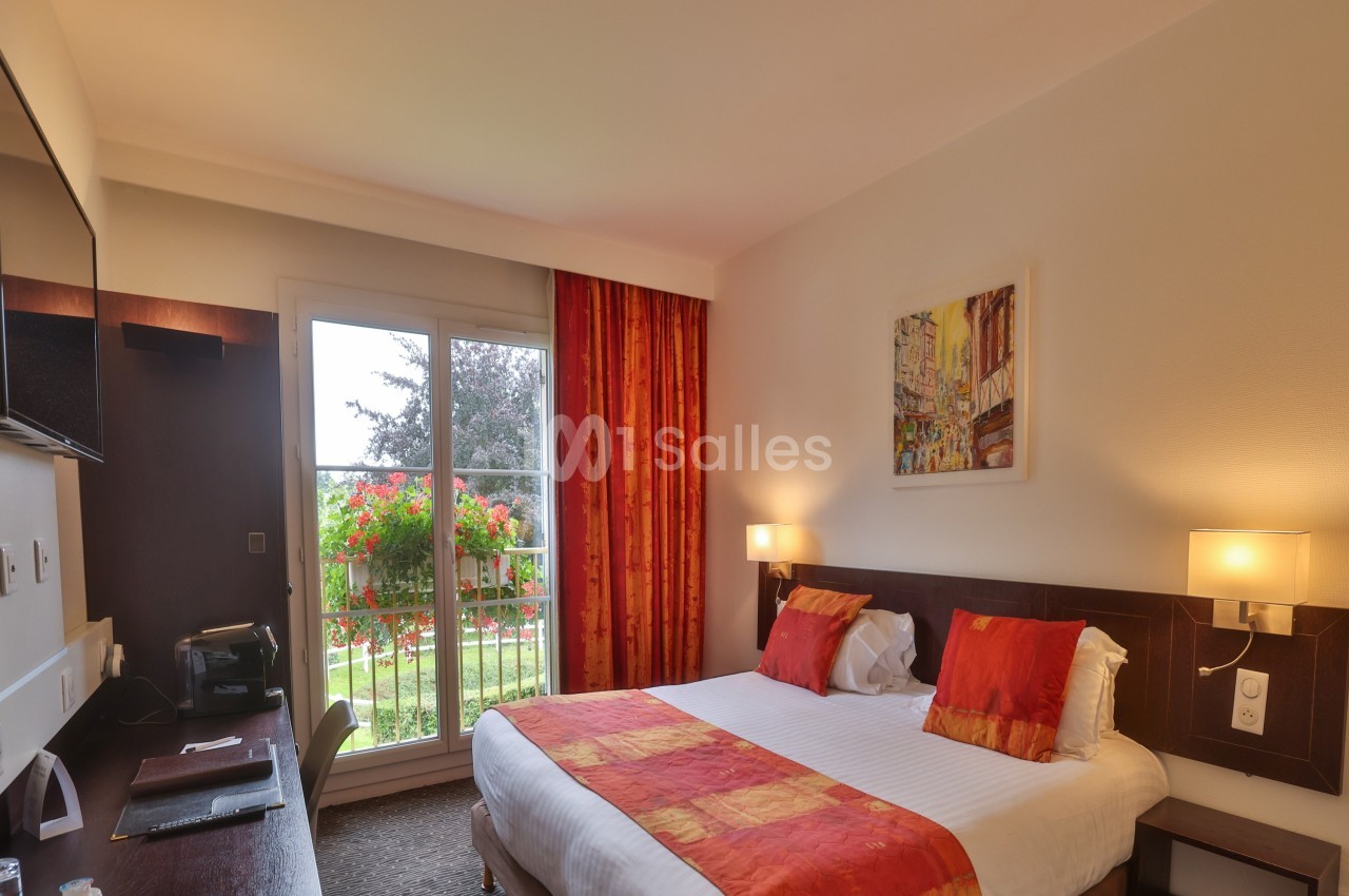 Chambre d'hôtel lumineuse avec lit double, bureau, télévision, et balcon donnant sur un jardin fleuri.