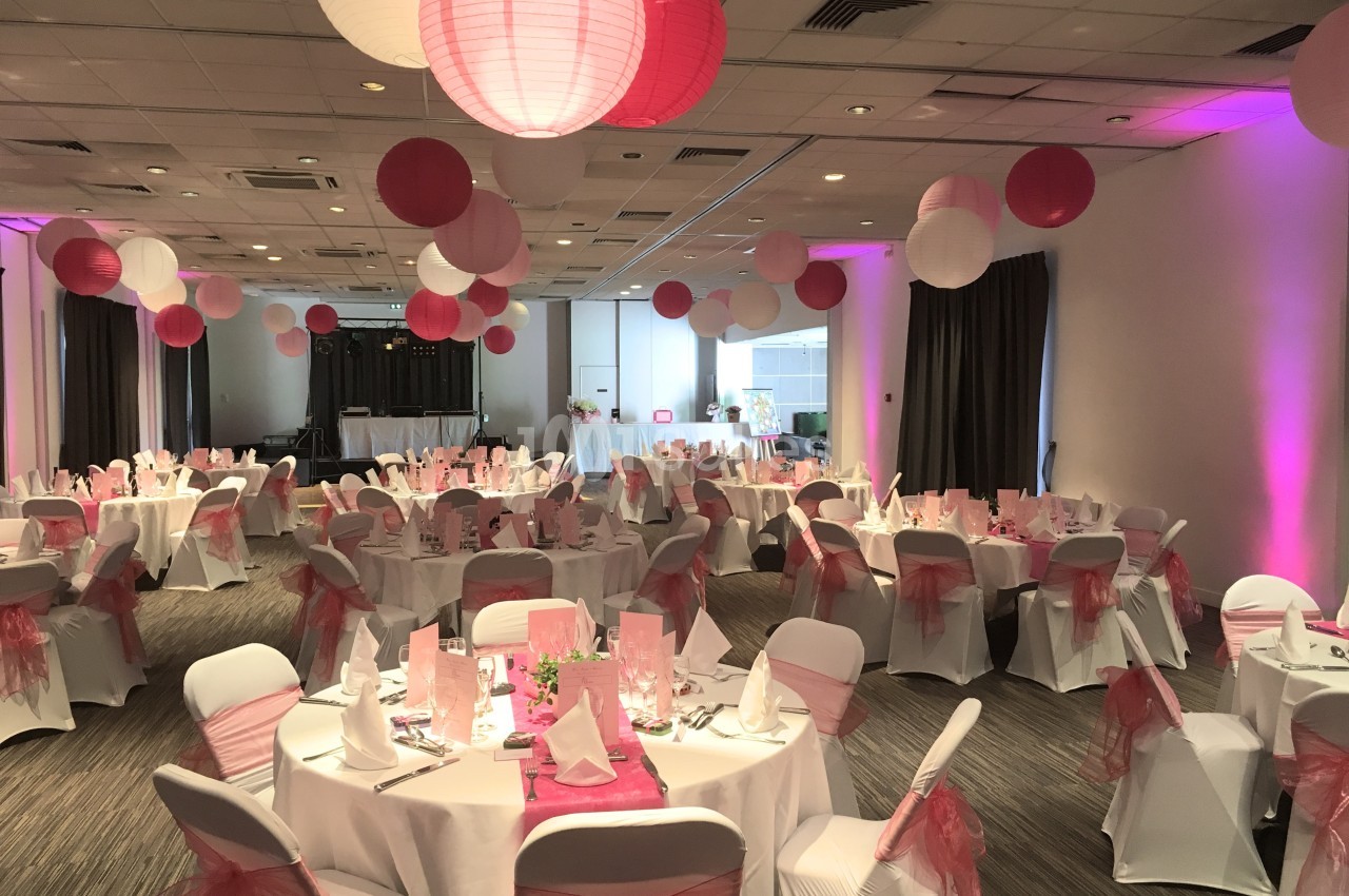 Salle de réception décorée avec des lanternes roses et blanches, tables rondes dressées et chaises ornées de nœuds roses.