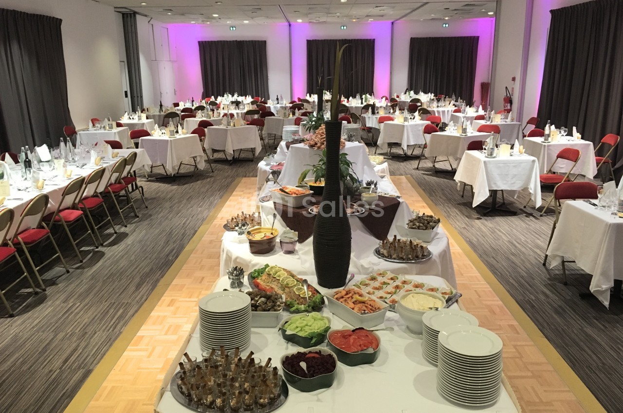 Salle de réception avec tables dressées et buffet central composé de plats variés, entouré de chaises rouges.