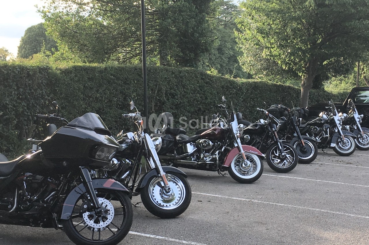 Plusieurs motos garées en ligne dans un parking extérieur bordé de haies et d'arbres.