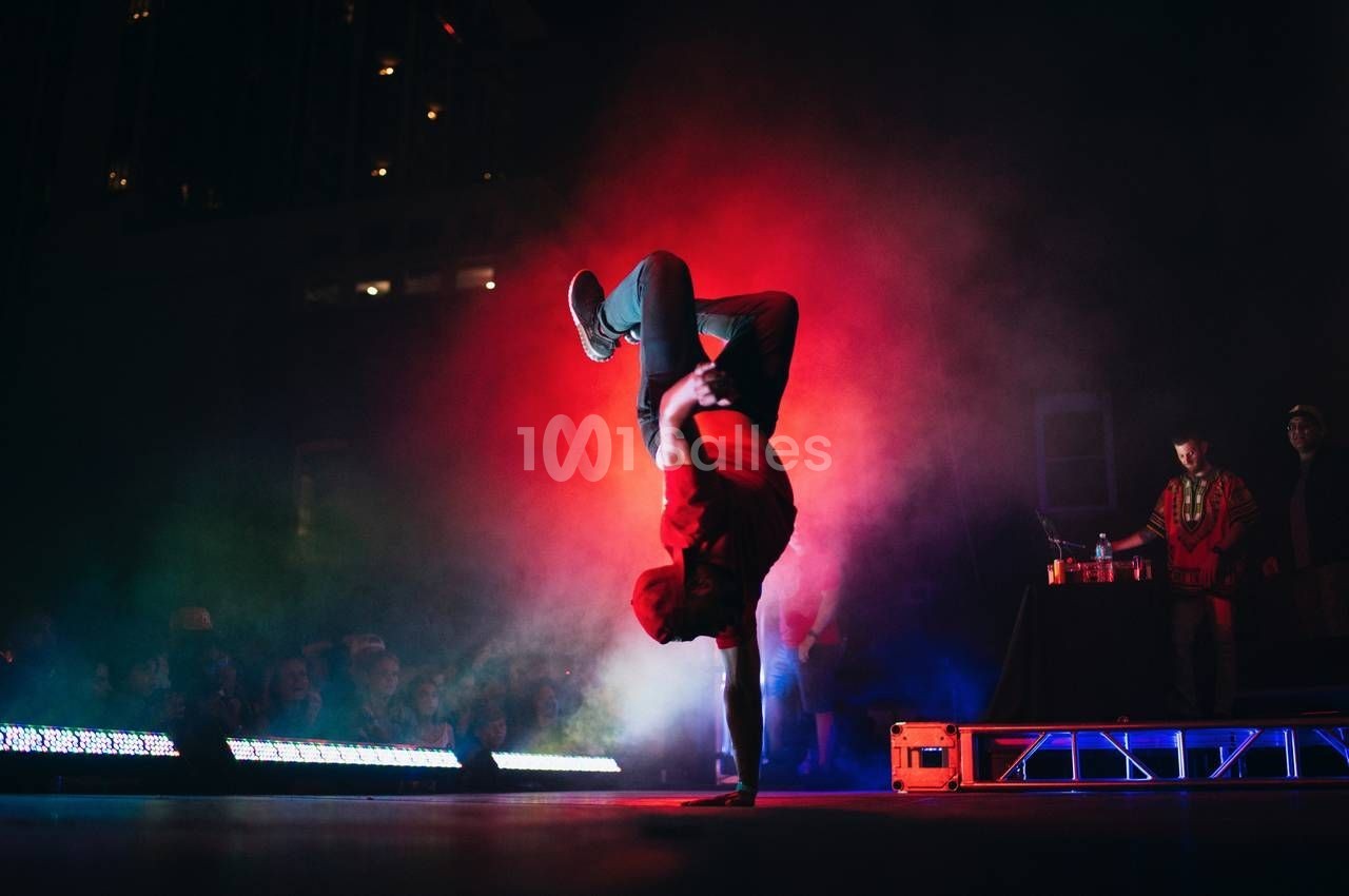 Un danseur effectue une figure acrobatique sur une main, éclairé par des lumières rouges et bleues sur une scène.