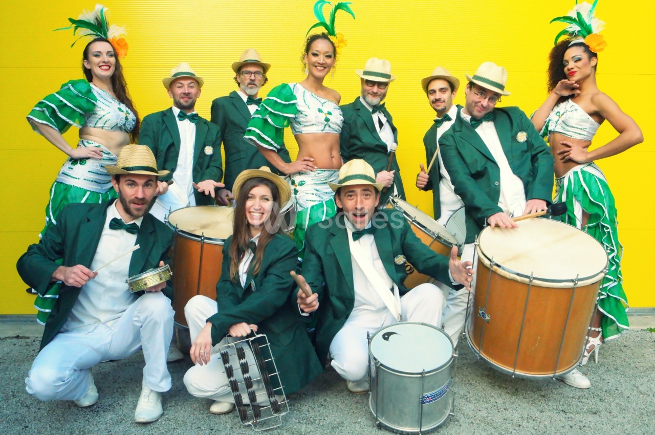 Un groupe de musiciens et danseuses en costumes verts et jaunes posant devant un mur jaune.