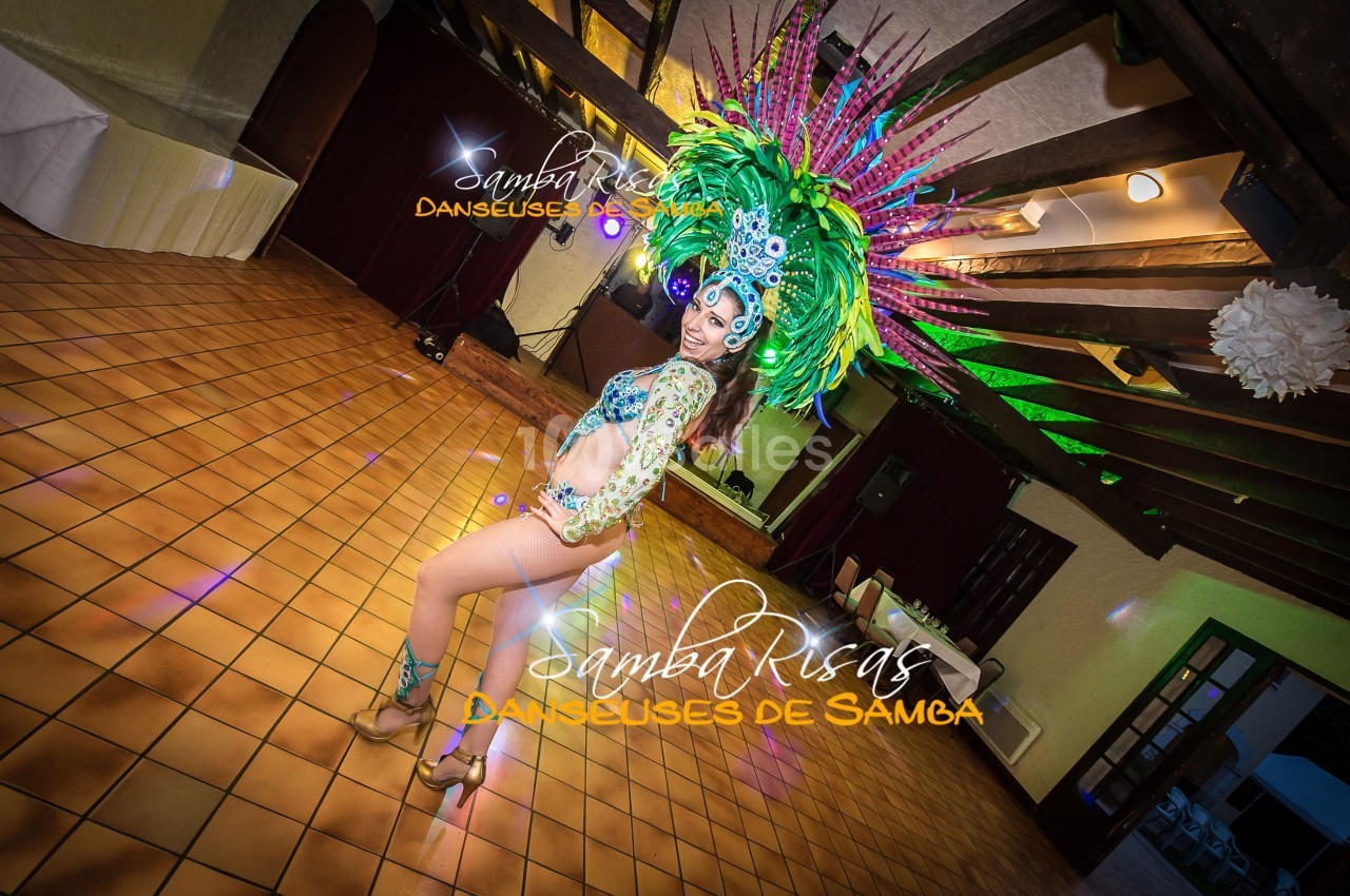 Danseuse de samba en tenue colorée avec une coiffe ornée de plumes, posant dans une salle au sol carrelé.