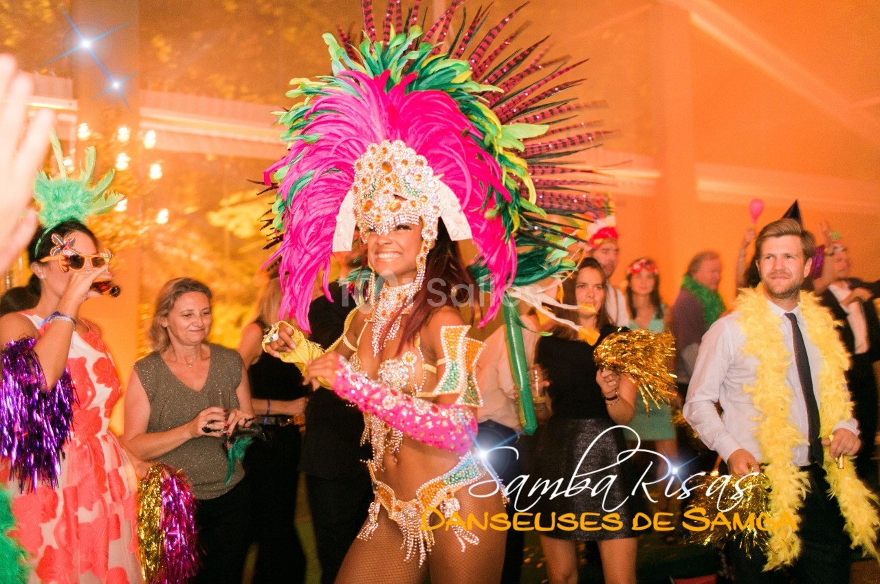 Danseuse de samba en costume coloré avec plumes, entourée de spectateurs lors d'une fête festive en intérieur.