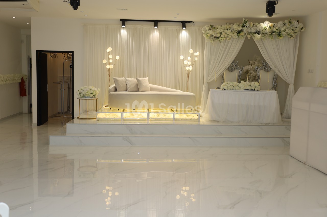 Salle lumineuse avec canapé blanc sur estrade, décorée de fleurs blanches et éclairages modernes.