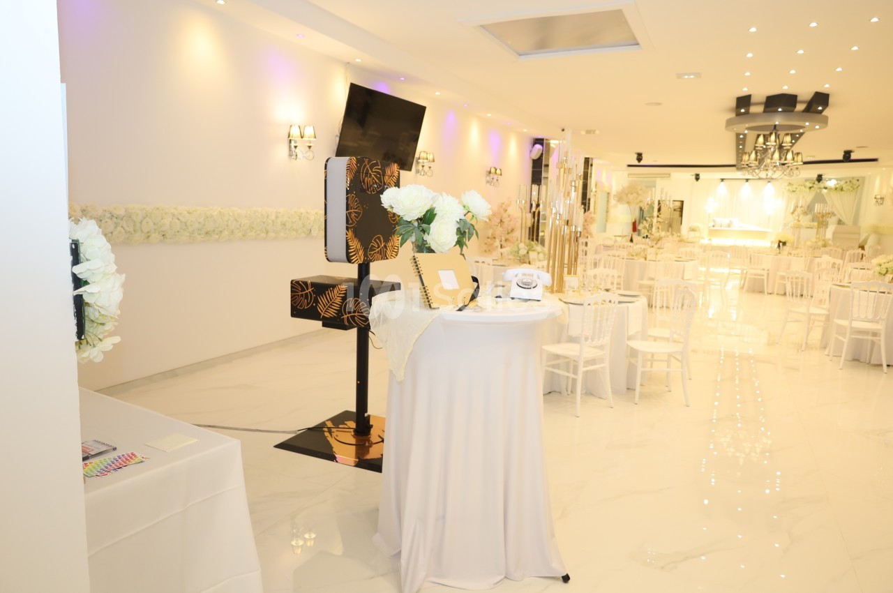 Salle de réception lumineuse avec tables décorées, fleurs blanches et un coin photo équipé d'accessoires.