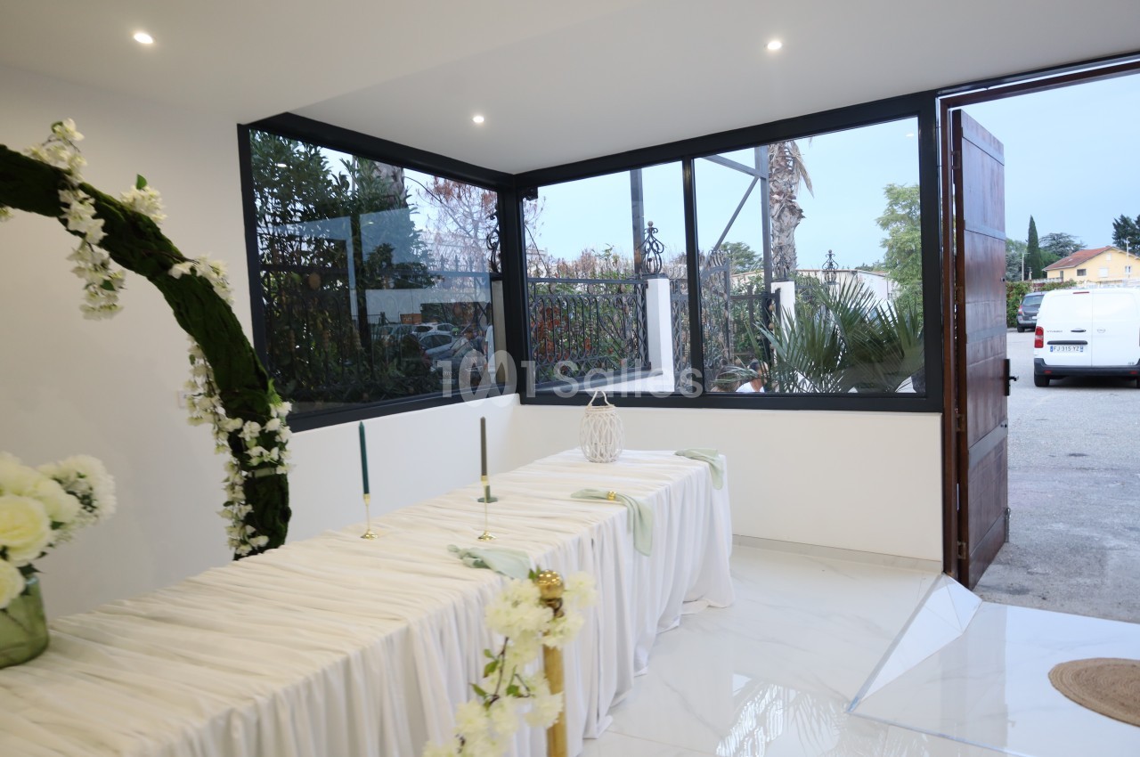 Salle lumineuse avec table drapée de blanc, décorée de fleurs et bougies, donnant sur un extérieur arboré.