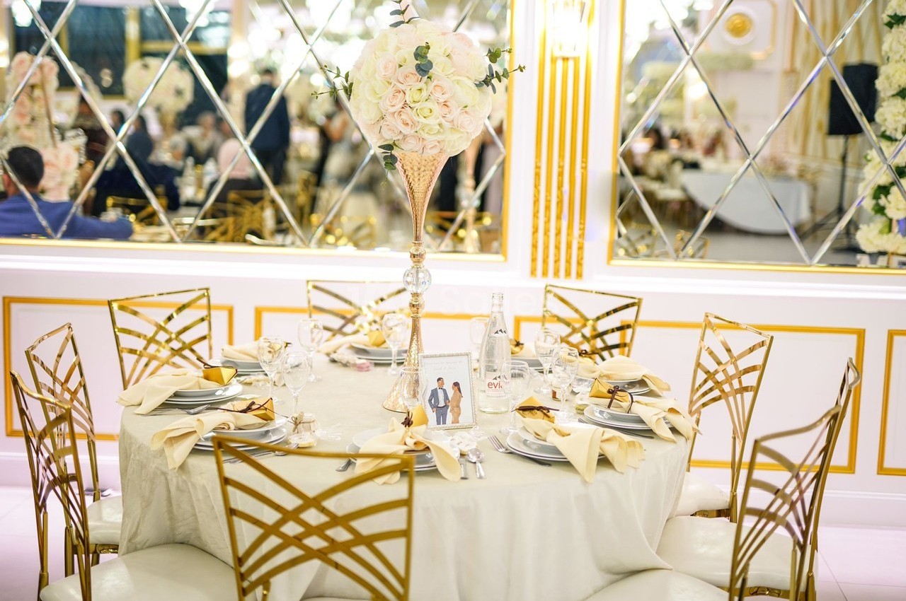 Table élégamment dressée avec nappes claires, chaises dorées et centre de table floral dans une salle ornée de miroirs.