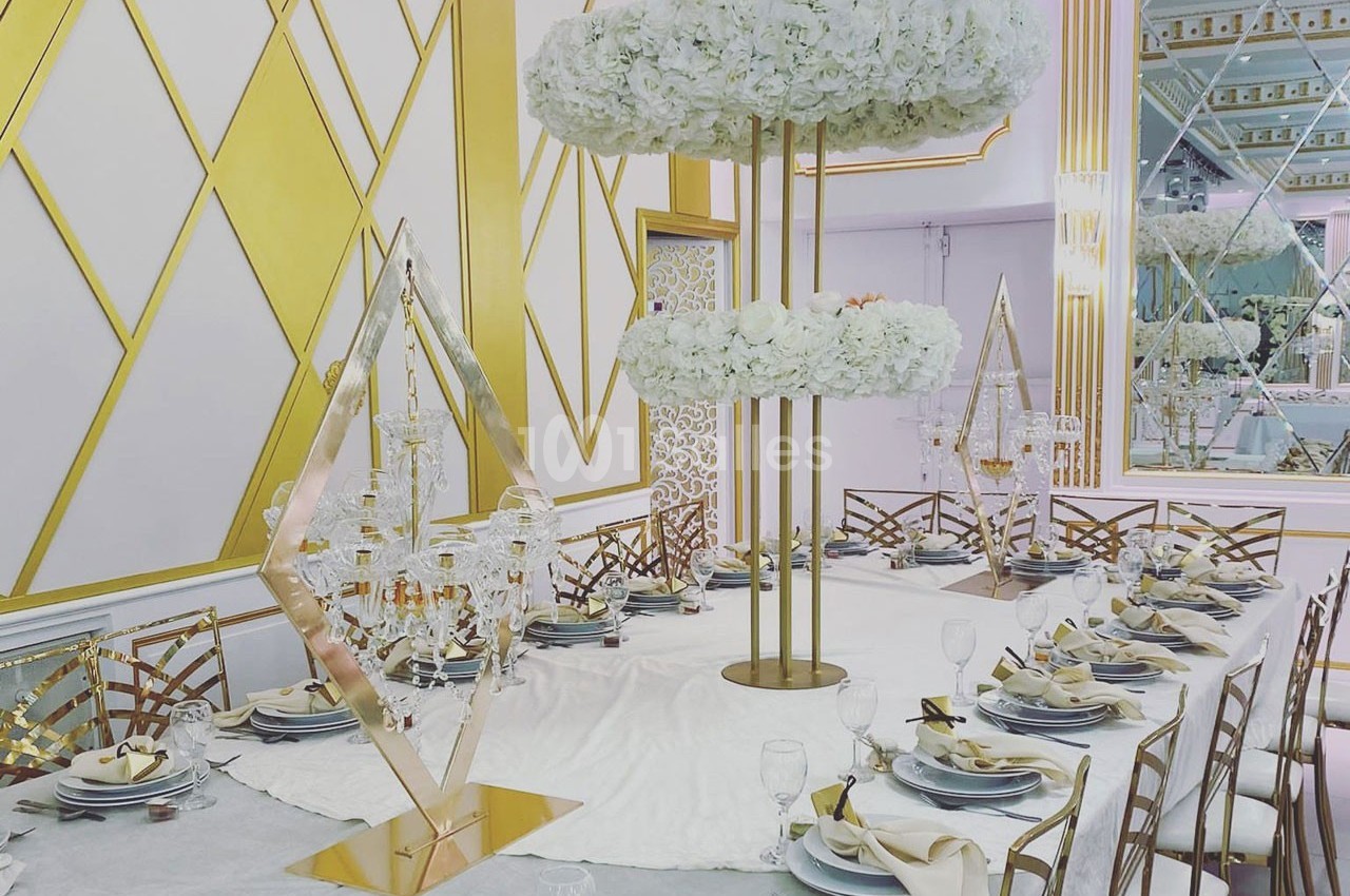 Table élégamment dressée avec nappes blanches, chaises dorées, décorations florales et éléments géométriques dorés.