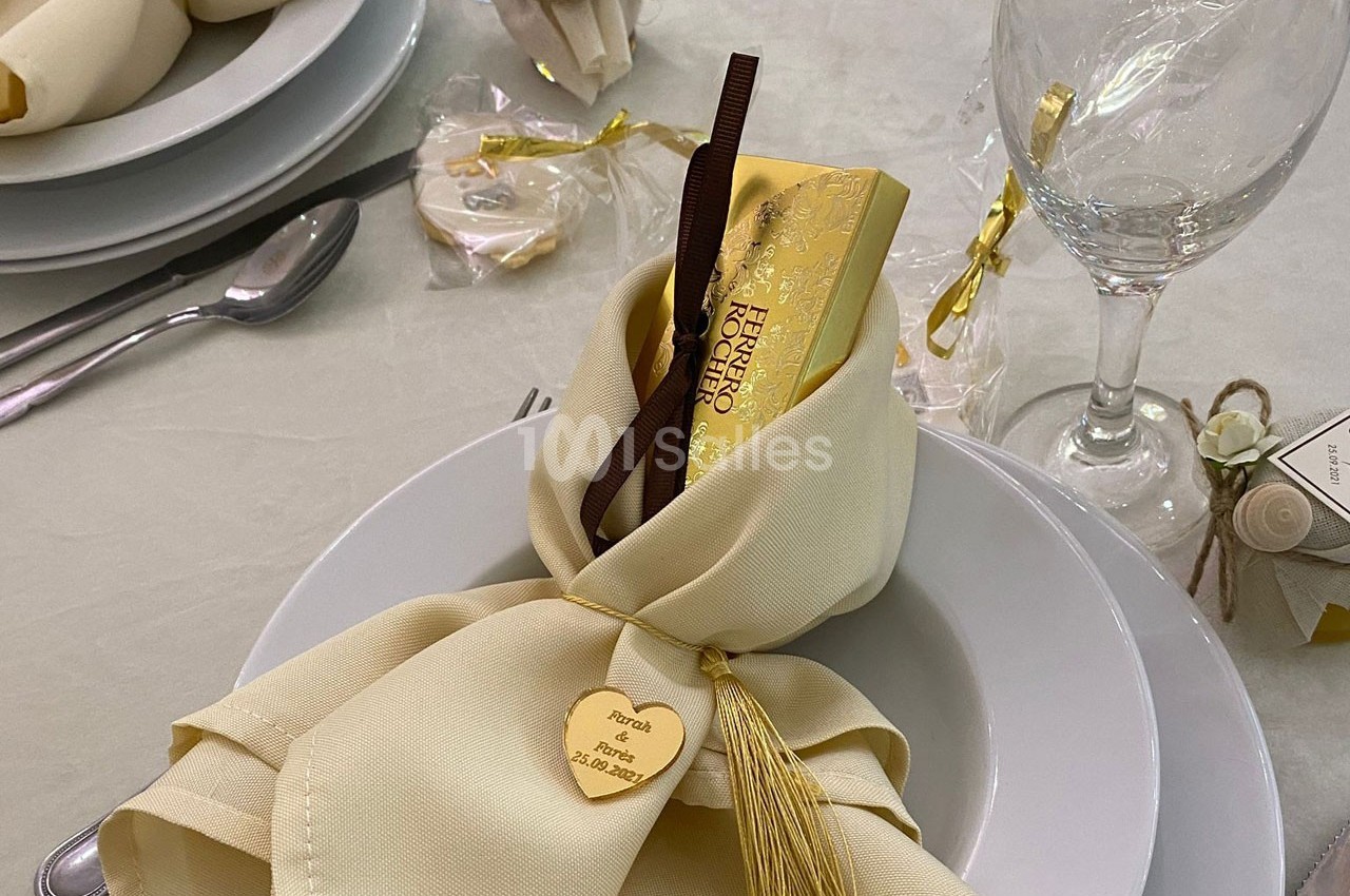Serviette beige pliée avec un ruban doré, contenant une barre de chocolat et un cœur gravé, posée sur une assiette blanche.