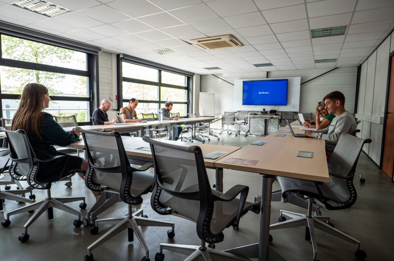 Salle de réunion lumineuse avec plusieurs personnes assises autour de tables, certaines utilisant des ordinateurs.