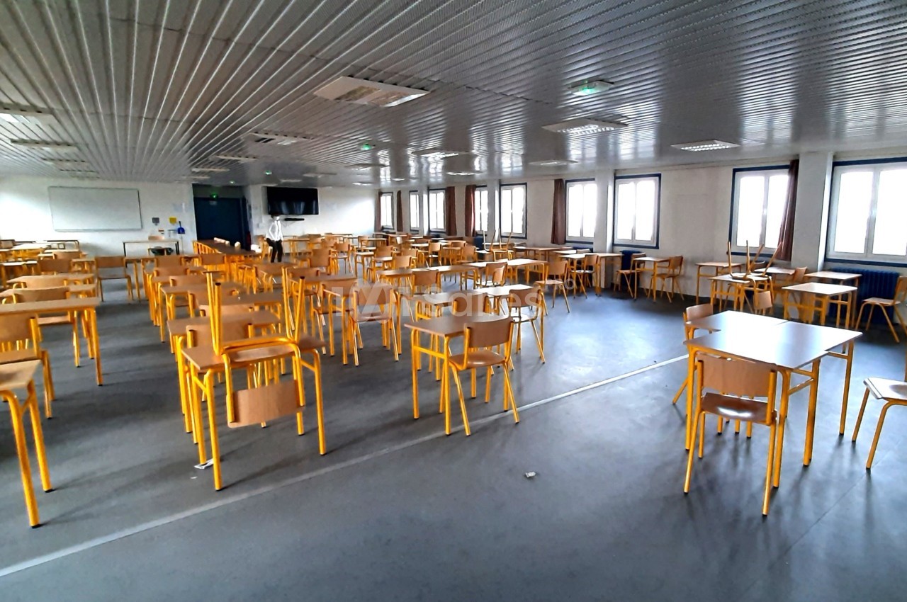 Salle de classe vide avec des tables et chaises jaunes alignées, éclairée par des fenêtres sur les côtés.