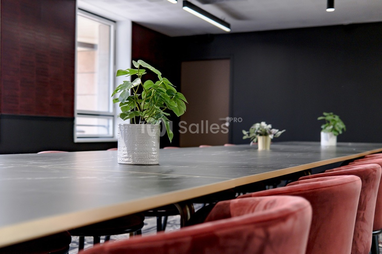Salle de réunion moderne avec une longue table noire, des chaises rouges et des plantes en décoration.
