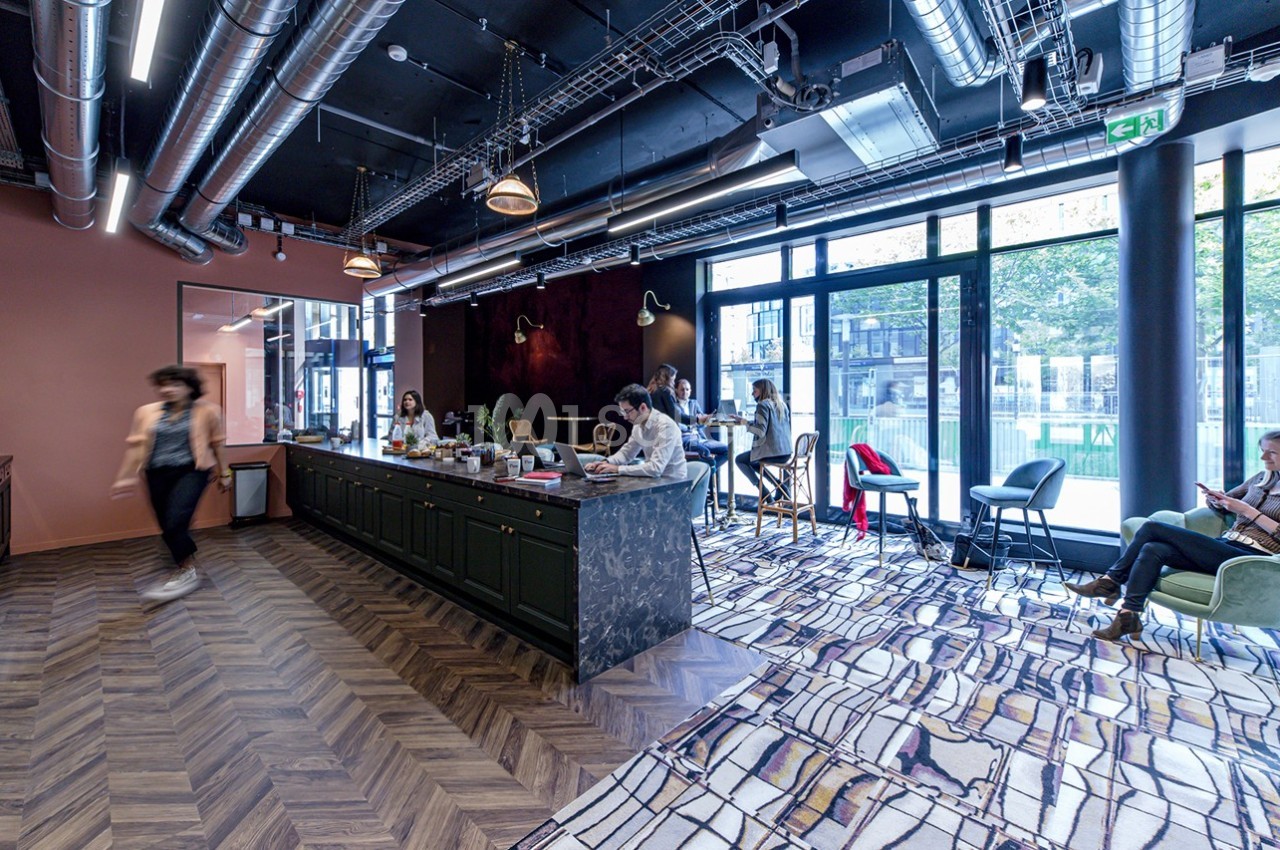 Espace de travail moderne avec cuisine ouverte, grandes baies vitrées et plusieurs personnes en activité.