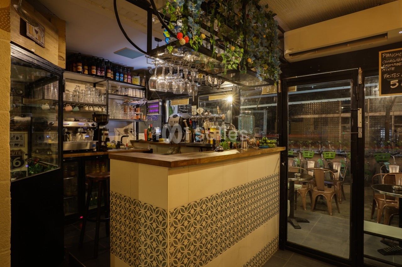 Comptoir de bar avec machine à café, verres suspendus et décoration végétale, donnant sur une terrasse extérieure.