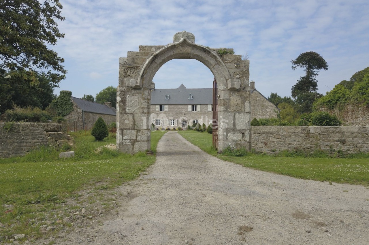 Portail en pierre ancienne encadrant une allée menant à une maison en pierre entourée de verdure.