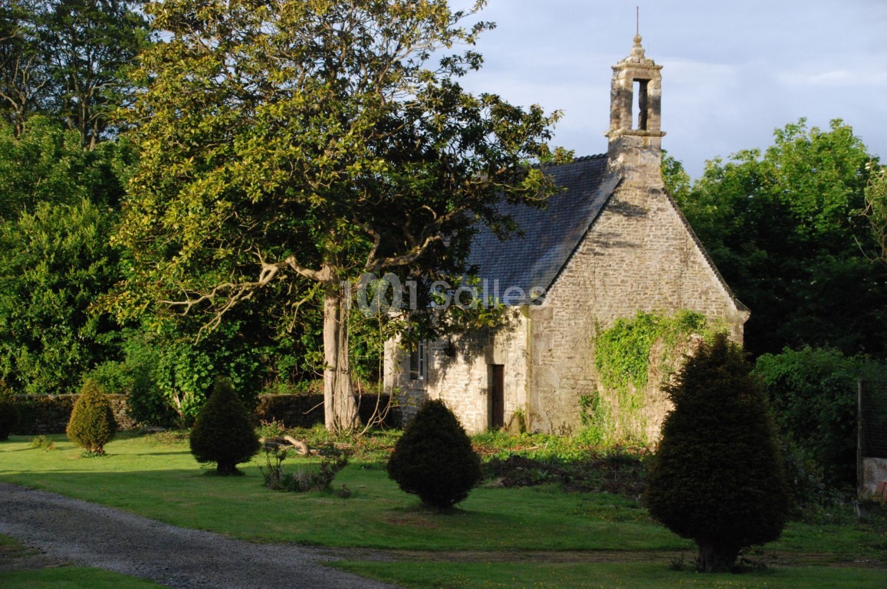 Petite chapelle en pierre entourée de verdure, avec un arbre imposant et des buissons dans un jardin.