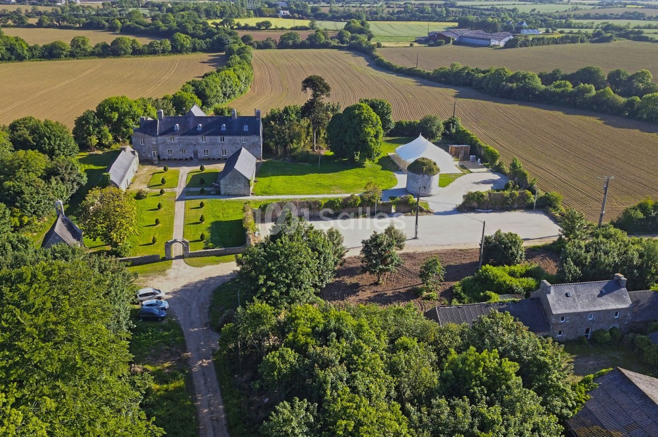 Vue aérienne d'un domaine rural avec bâtiments en pierre, jardin aménagé, moulin et champs environnants.