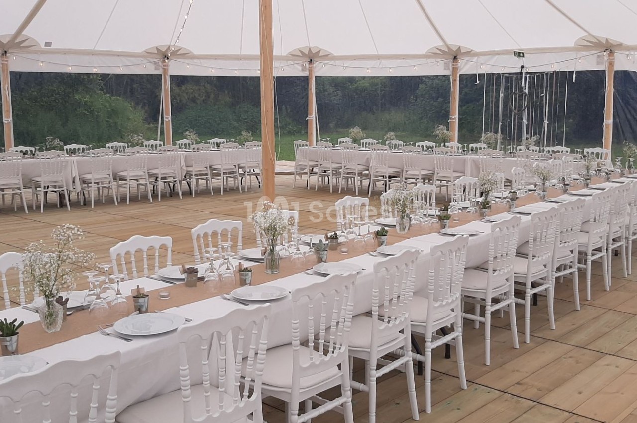 Tables longues décorées de nappes blanches et chaises assorties sous une tente ouverte, prêtes pour un événement.