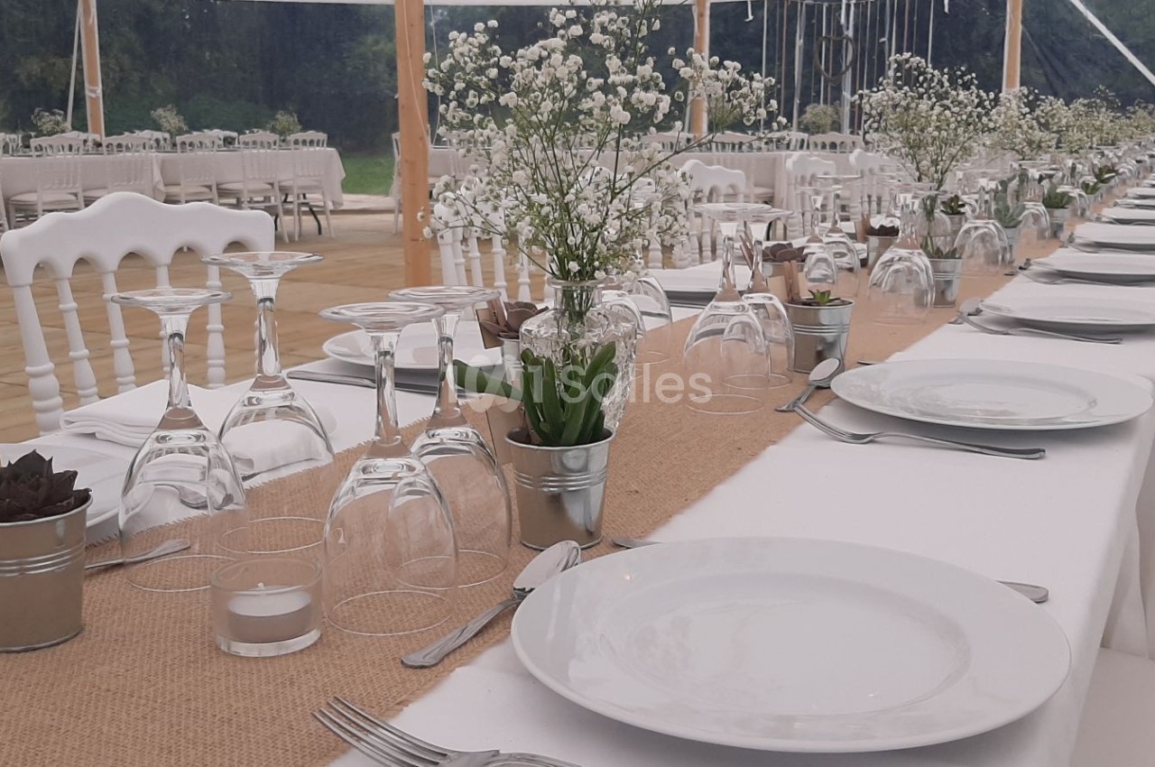 Tables de réception alignées avec nappes blanches, vaisselle, verres, et décorations florales dans un cadre extérieur.