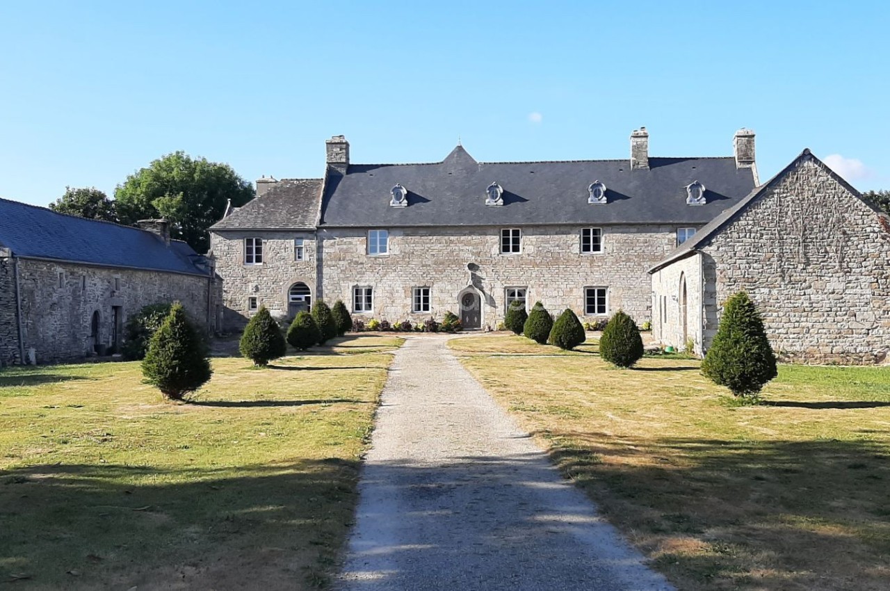 Manoir en pierre entouré de pelouses, avec une allée centrale bordée de petits arbustes taillés.