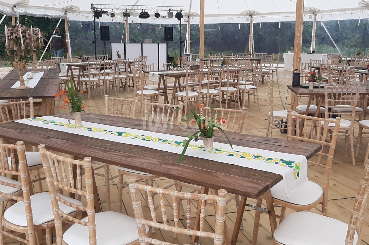 Salle de réception sous chapiteau avec tables en bois, chaises en bois clair et décorations florales.
